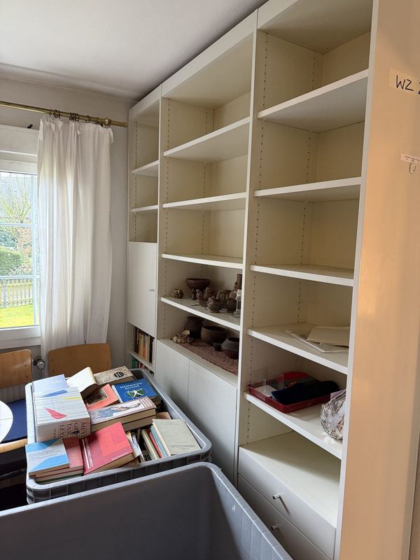 Fast geleertes Bücherregal Weißes Bücherregal, bis auf einige Gegenstände vollständig geleert, vor dem Regal ein grauer Rollbehälter mit Büchern