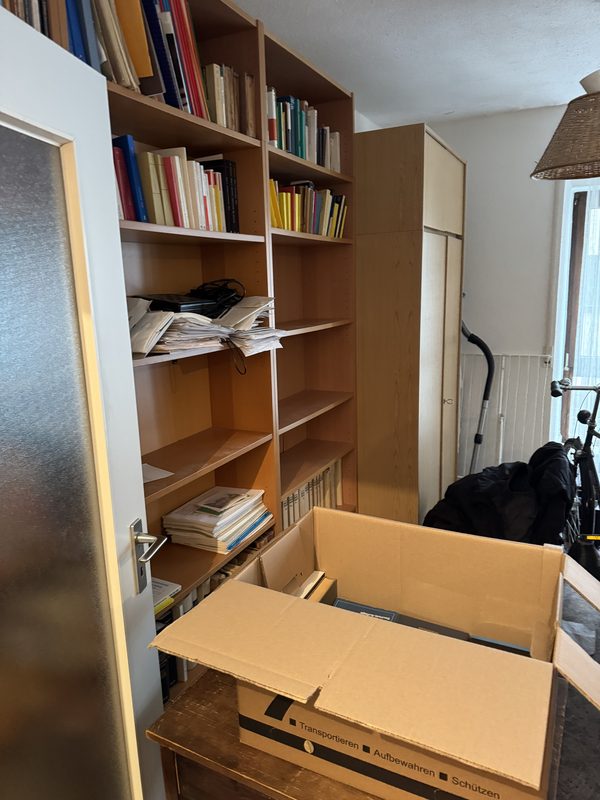 Ein offener, noch leerer Umzugskarton vor einem Regal, in dem Bücher und Papierstapel liegen