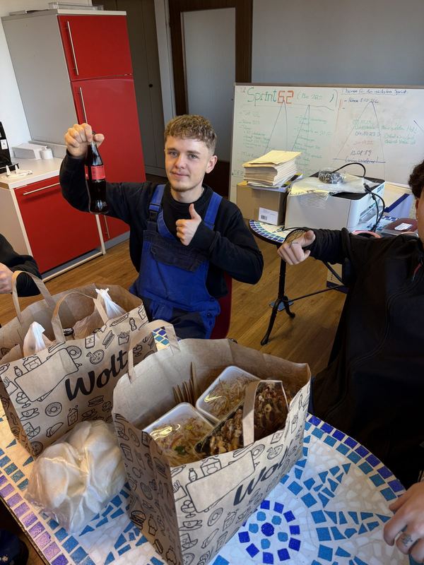 Mit Getränk in der Hand und erhobenem Daumen sitzt ein Mitarbeiter vor dem gemeinsamen Mittagessen