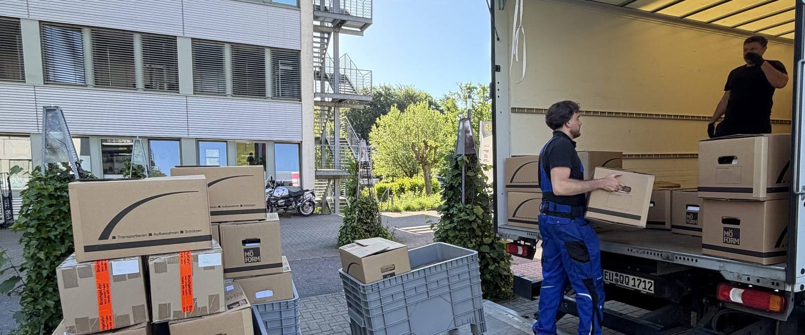 Einblicke in unseren Umzugsalltag in Bonn: Vor einem Bürogebäude mit Außentreppe steht ein Umzugswagen mit offenem Laderaum, ein Mitarbeiter trägt einen Karton zur Ladebordwand, ein zweiter steht im Laderaum, im Vordergrund gestapelte Umzugskartons und graue Kunststoffboxen.