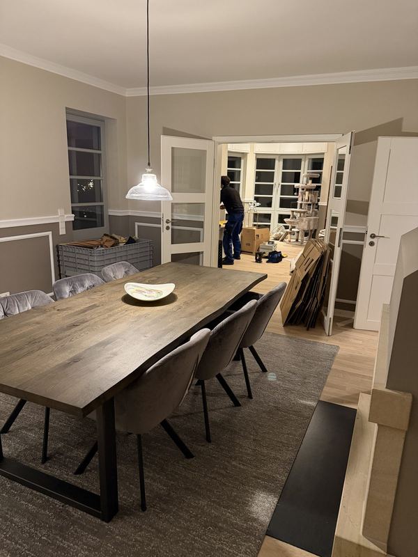 Fertig eingerichtetes Esszimmer, der Tisch ist gedeckt, nebenan werden leere Kartons zusammengefaltet
