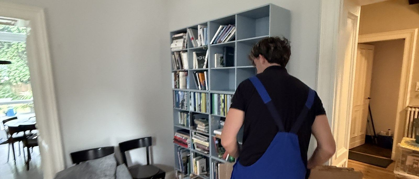 Fragen und Antworten zum Umzug in Bonn: Ein Mitarbeiter in Latzhose steht vor einem hellgrauen Würfelregal mit Büchern und Ordnern und packt dessen Inhalt in einen Karton, im Vordergrund ein Esstisch mit Stühlen.