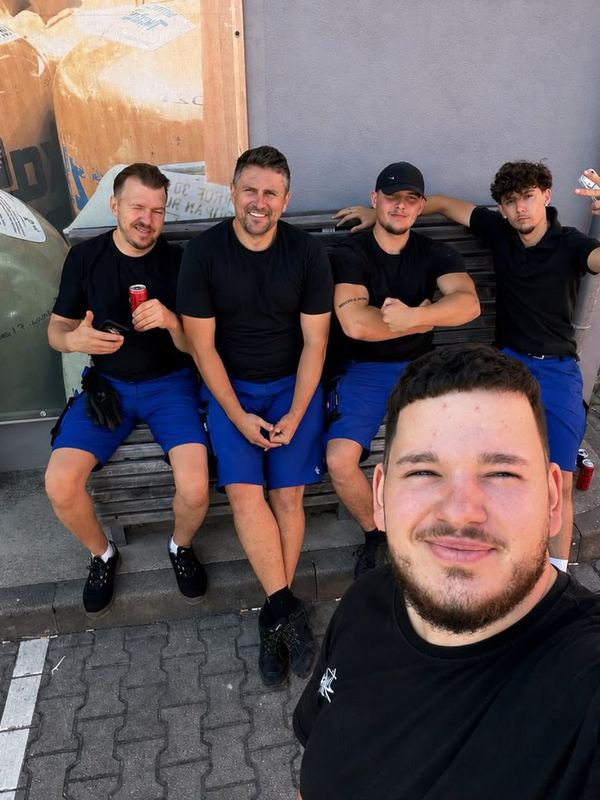 In der Sonne sitzen fünf Mitarbeiter in Arbeitskleidung gemeinsam auf einer Bank