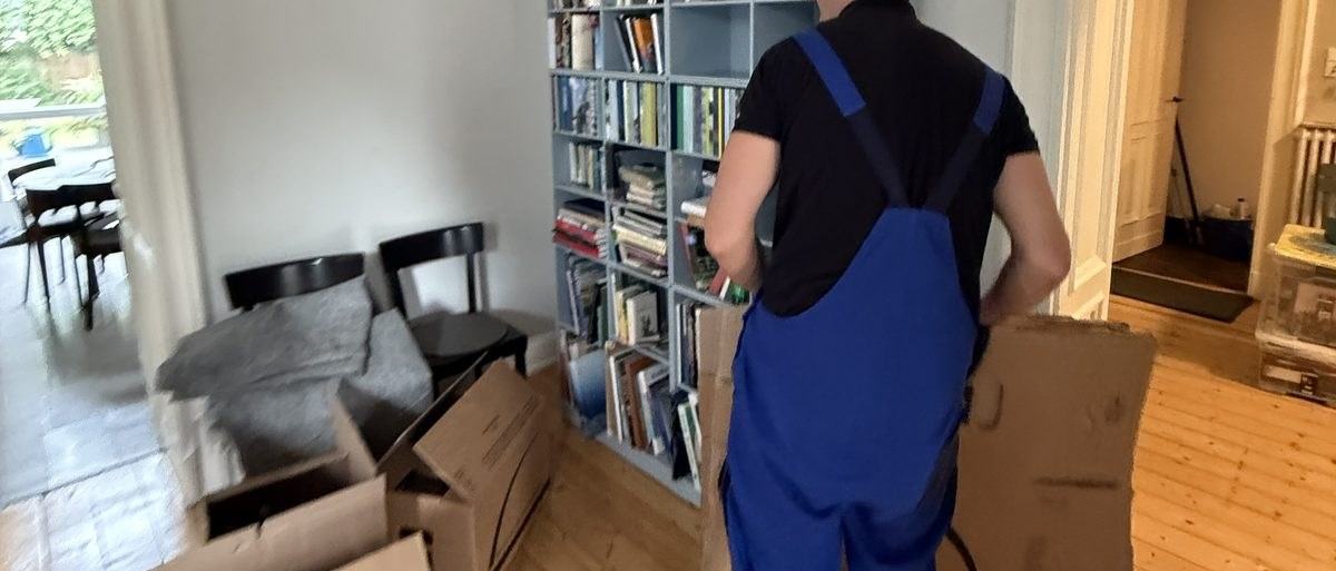 Haushaltsauflösung in Bonn: Ein Mitarbeiter in Latzhose geht mit einem gefalteten Karton an einem gut gefüllten Bücherregal vorbei, im Vordergrund Stühle und weitere Kartons