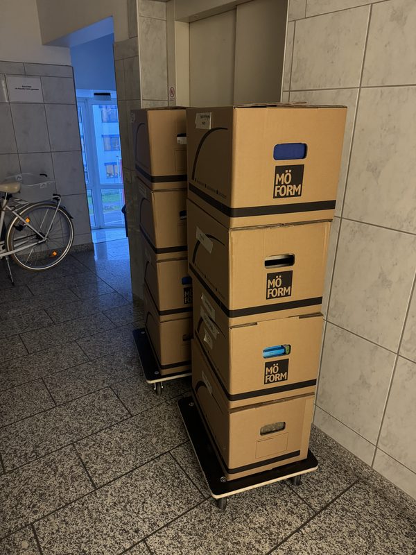 Ein Stapel beschrifteter Kartons steht auf einem Transportroller in einem Hausflur mit Fliesenboden, bei einem Umzug in Bonn