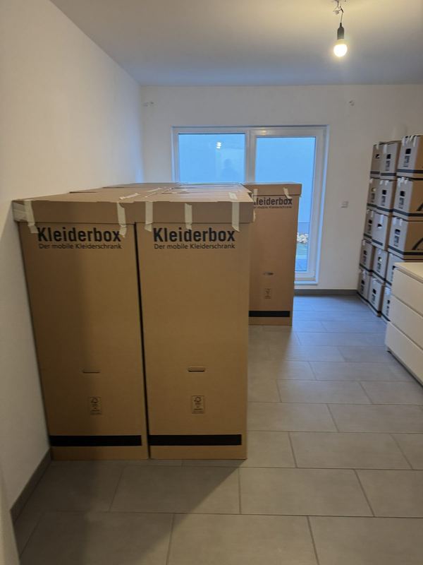 In einem leeren Zimmer stehen zwei Kleiderboxen neben gestapelten Kartons