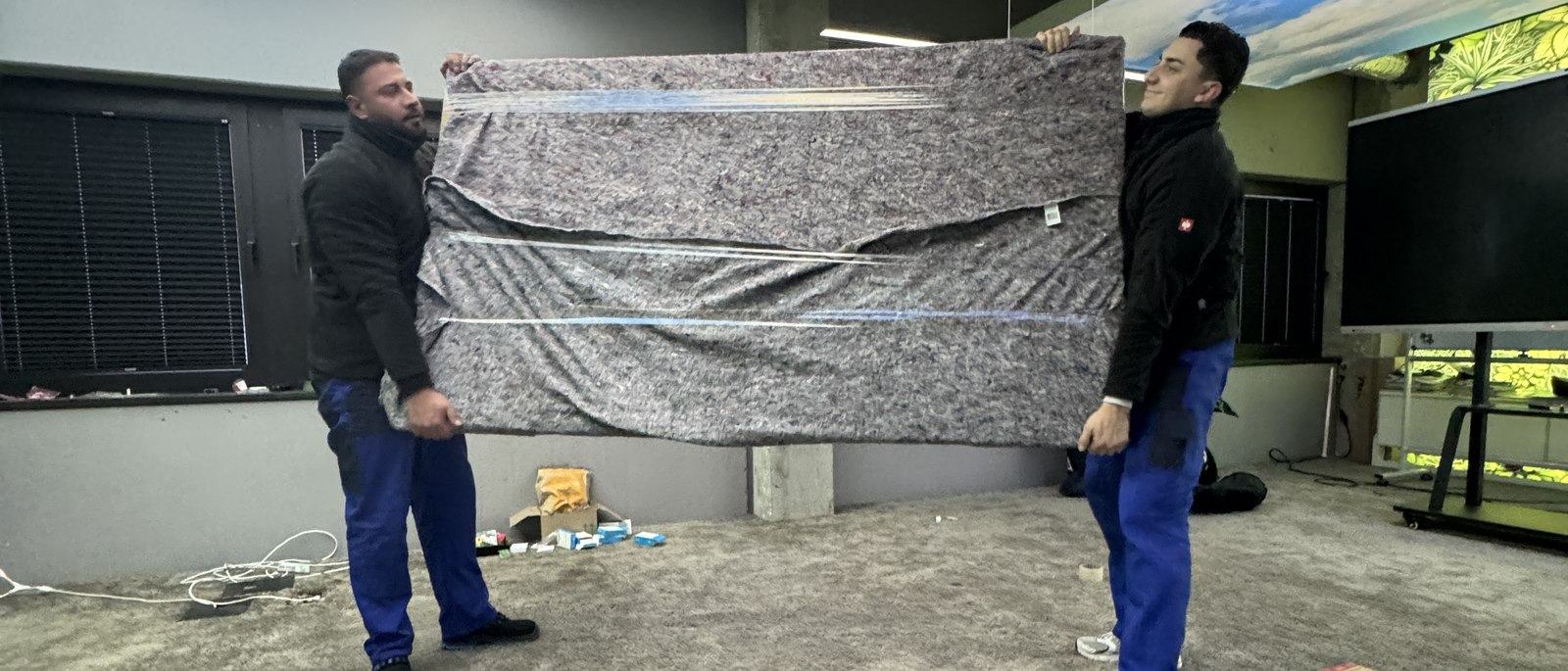Ihr Kontakt in Bonn: Zwei Mitarbeiter tragen zu zweit ein großes flaches Möbelstück, das in eine graue Umzugsdecke gewickelt ist, über einen Hof an einer Hauswand entlang.