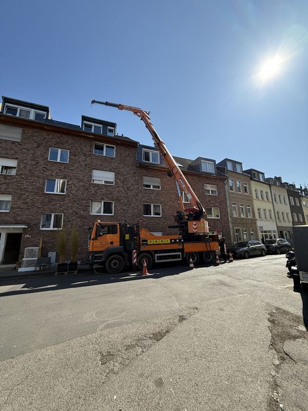 In einer Wohnstraße steht ein orangefarbener Kranwagen mit ausgefahrenem Ausleger, davor Warnkegel