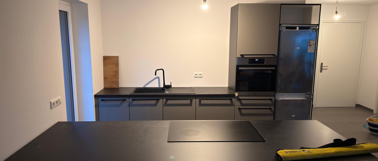 Küchenmontage in Bonn: Eine fertig montierte moderne Küche mit dunklen Fronten, Kochinsel mit Induktionsfeld, Spüle, Backofen und Kühlschrank in einem hellen leeren Raum, auf dem Boden liegt noch eine Werkzeugtasche.