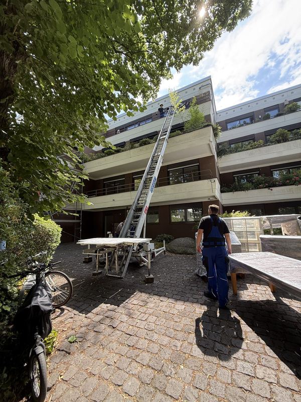 An einem mehrgeschossigen Wohnhaus steht ein ausgefahrener Möbellift, unten wartet ein Mitarbeiter