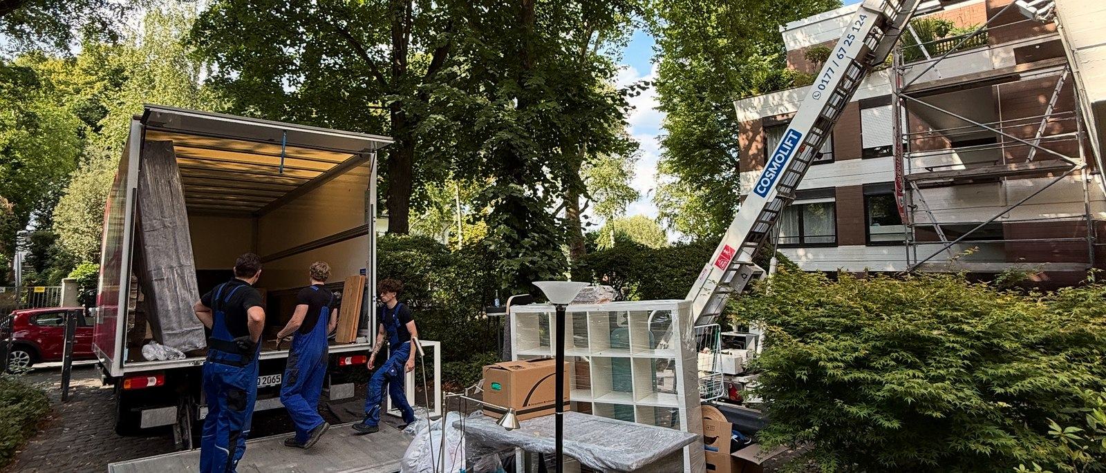 Möbellift für Umzüge in Bonn: Ein ausgefahrener Möbellift ragt bis zum obersten Geschoss eines eingerüsteten Wohnhauses, unten stehen drei Mitarbeiter am geöffneten Laderaum eines Lkw neben einem Regal und Kartons