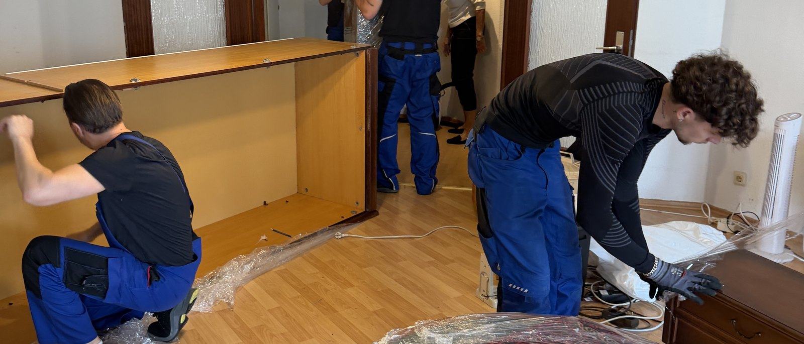 Möbelmontage in Bonn: Drei Mitarbeiter zerlegen in einem Wohnzimmer mit Parkett eine große Schrankwand aus Holz, die Seitenteile liegen bereits am Boden, daneben Folie und Werkzeug.