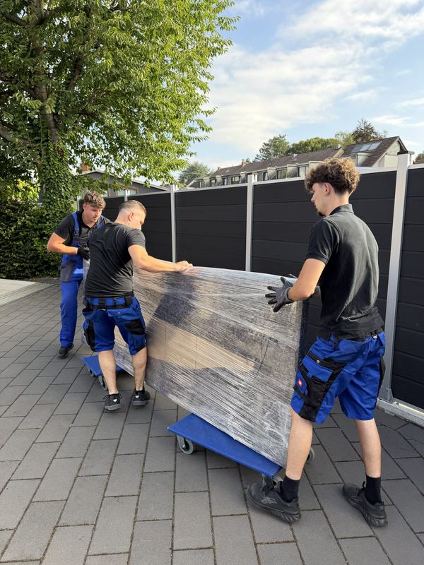 Über eine Einfahrt rollen drei Mitarbeiter ein in Folie gepacktes Möbelstück auf Rollbrettern zum Fahrzeug