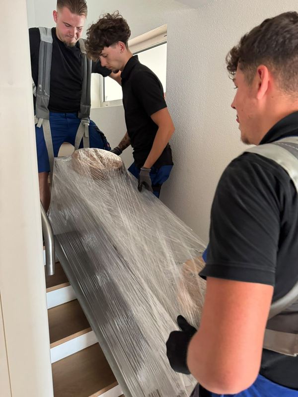 Zu dritt bringen Mitarbeiter ein in Folie gepacktes Möbelstück die Treppe hinab