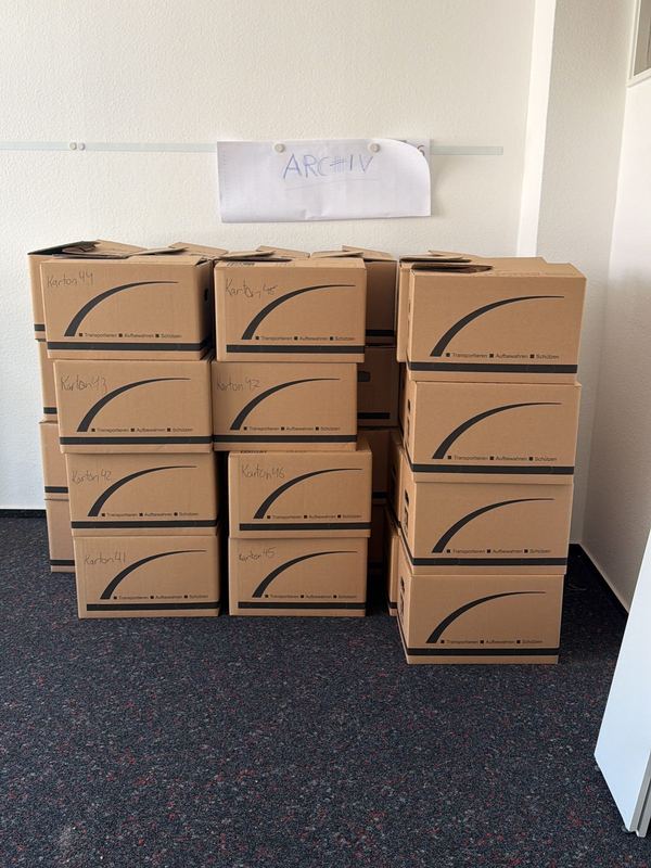 Unter einem handgeschriebenen Raumschild stapeln sich nummerierte Umzugskartons in einem leeren Büro