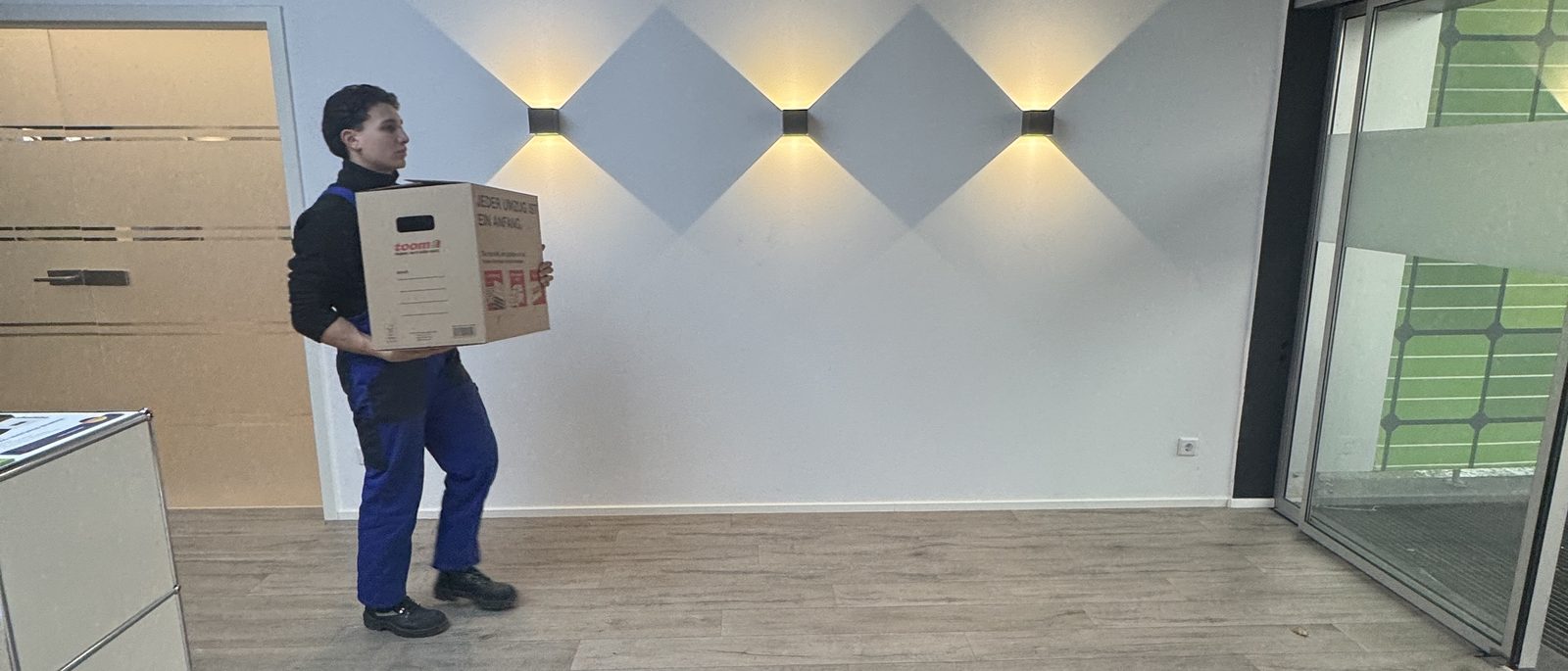 Praxisumzug in Bonn: Ein Umzugshelfer in blauer Arbeitshose trägt einen Karton an einer hellen Wand mit drei Wandleuchten vorbei