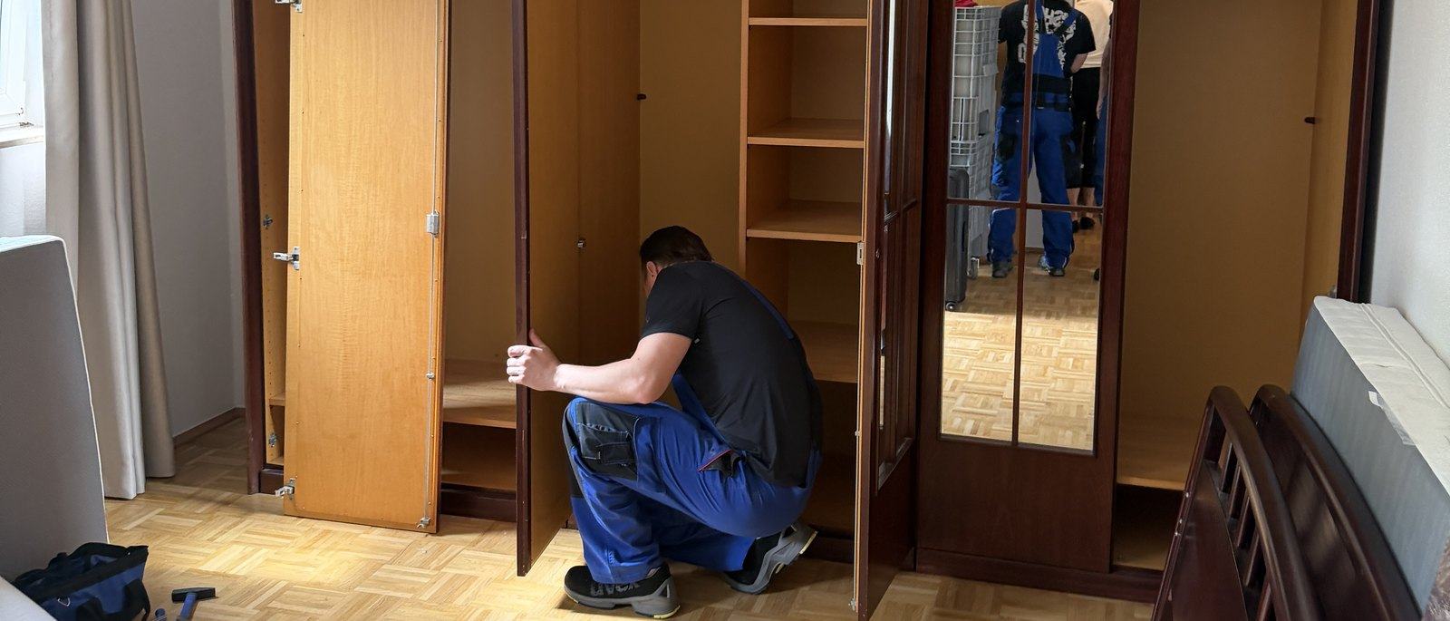 Seniorenumzug in Bonn: Vor einem geleerten Kleiderschrank aus dunklem Holz kniet ein Mitarbeiter, das Schlafzimmer hat Parkettboden