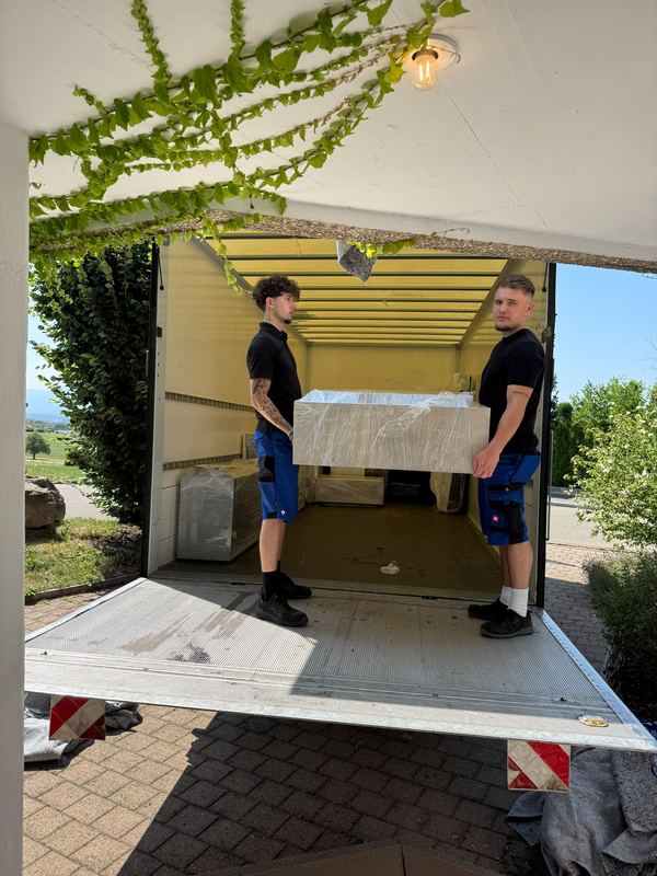 Verpacktes Möbelstück über die Ladebordwand laden Zwei Kollegen hieven ein foliertes Möbelstück an der Ladebordwand hoch in den Laderaum des Umzugswagens