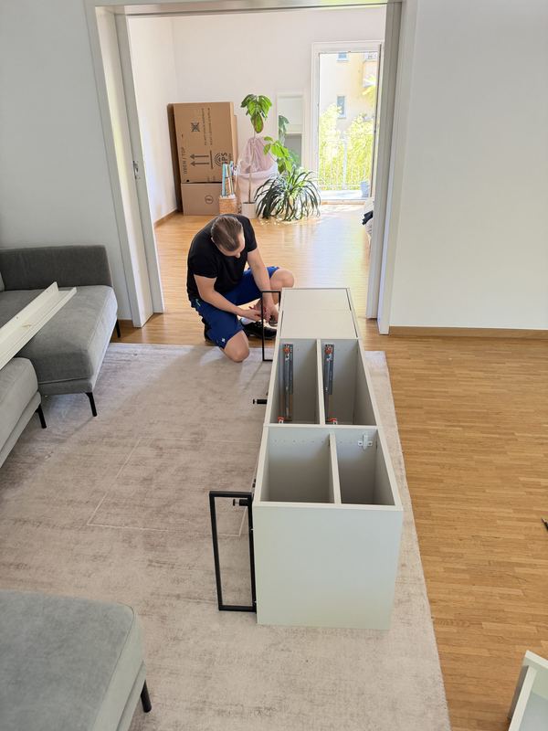 Sideboard am neuen Wohnort aufbauen Ein Kollege kniet vor einem weißen Sideboard in einer hellen Wohnstube und montiert es, hinter ihm stapeln sich Umzugskartons