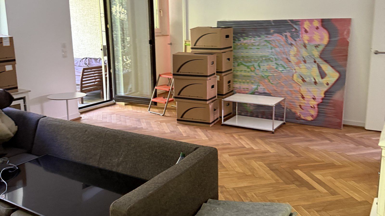 Umzugscheckliste für Bonn: Schritt für Schritt zum neuen Zuhause: In einem Wohnzimmer mit Fischgrätparkett stehen gestapelte Umzugskartons neben einem Sofa und einem Beistelltisch, an der Wand lehnt ein großes buntes Bild, dahinter eine Terrassentür.