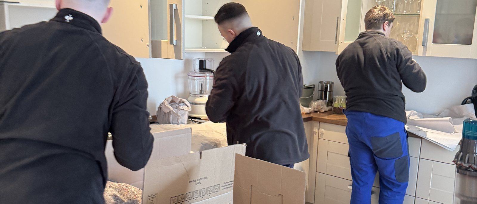 Umzugskartons und Kleiderboxen für Ihren Umzug in Bonn: Drei Mitarbeiter packen in einer Küche mit hellen Fronten Geschirr und Kleingeräte in Umzugskartons auf der Arbeitsplatte, in der Mitte ein offener Karton mit Packpapier.
