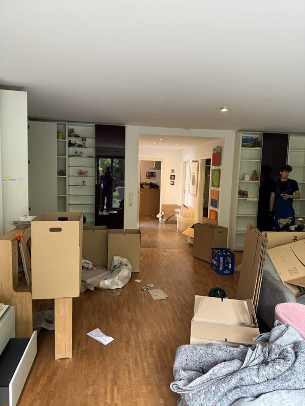 Wohnzimmer mit schon leeren Einbauregalen und bereitgestellten Kartons, im Hintergrund zwei Mitarbeiter